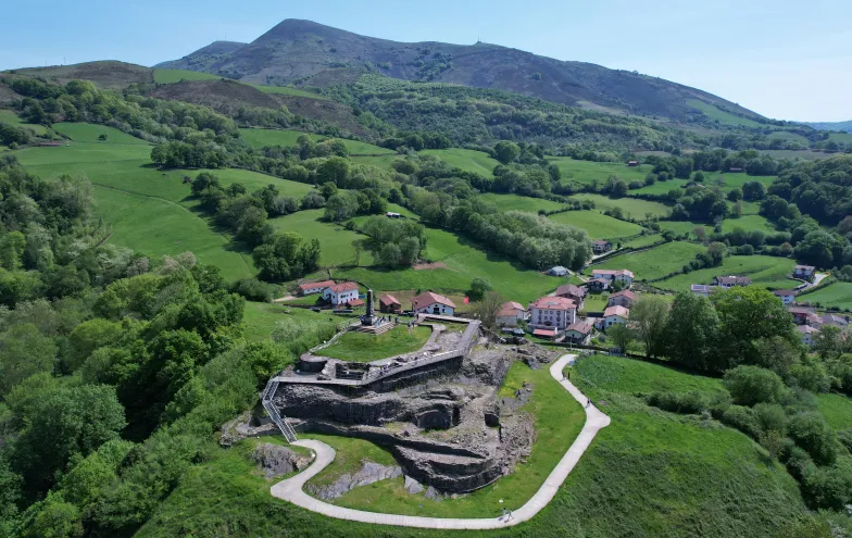 Castillo de Amaiur en valle verde de Baztan rodeado de bosque, praderas y pueblo cercano con montaña al fondo.