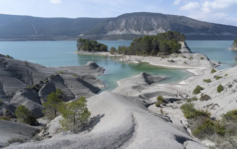 Embalse de Yesa con aguas turquesas, formaciones rocosas grises y vegetación dispersa, montañas al fondo.