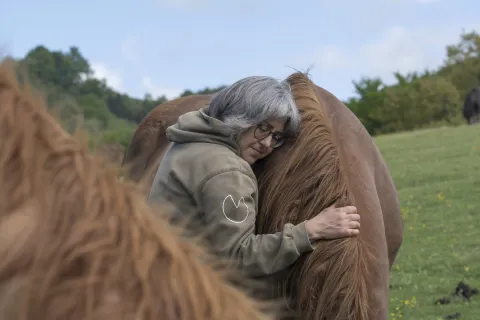 Propietaria de la experiencia de Caballos del bosque abrazando caballo marrón con paisaje de montaña verde al fondo.