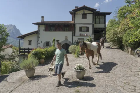 Uno de los propietarios de la experiencia Ekino Aukera paseando caballos con una casa rural tradicional detrás.