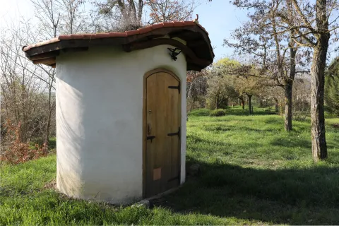 Pequeña caseta de piedra blanca con tejado de barro en Lizoain-Arriasgoiti, rodeada de árboles y prado.