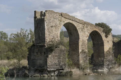 Ruinas de puente medieval con arcos de piedra sobre río en Puente de los Roncaleses, rodeado de vegetación y montaña.