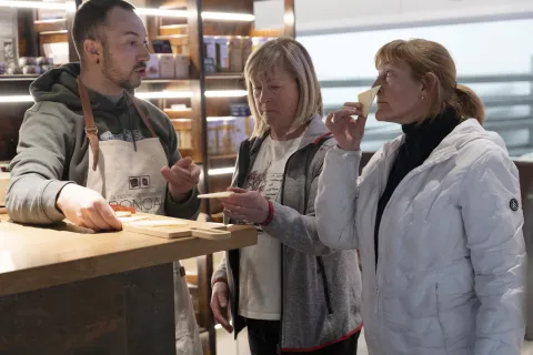 Artesano en Quesos Ekia explica elaboración de queso a visitantes en tienda con productos locales al fondo.