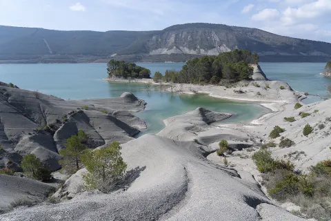 Embalse de Yesa con aguas turquesas, formaciones rocosas grises y vegetación dispersa, montañas al fondo.