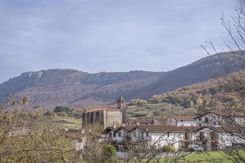 Iglesia con campanario sobre pueblo de Urdiain, rodeado de montañas boscosas y prados verdes.