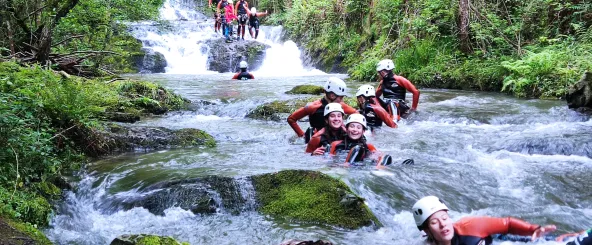 Descenso de cañón en río con participantes en traje de neopreno rojo y casco blanco, bosque atlántico denso y cascada de fondo.