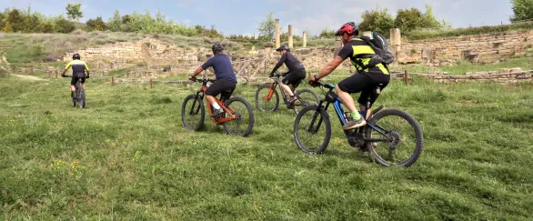Grupo de ciclistas en prado cerca de ruinas romanas en Territorio Eslava, patrimonio arqueológico al fondo.
