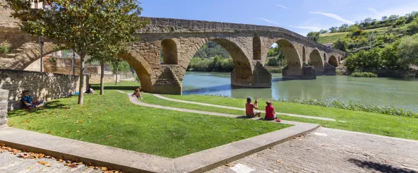 Dos personas sentados en la orilla observando el puente de los peregrinos en Puente la Reina