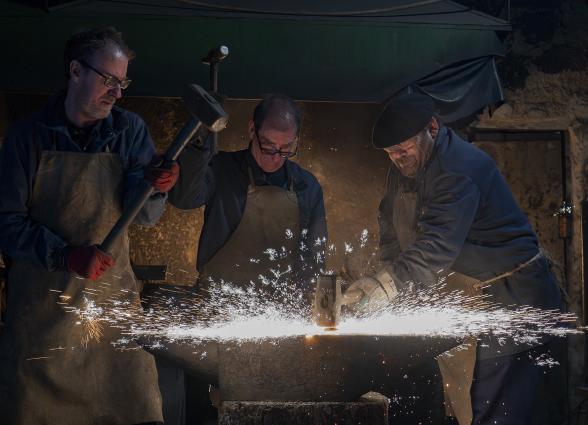 Tres herreros en taller de forja tradicional trabajando el metal con martillo y yunque, produciendo chispas incandescentes.