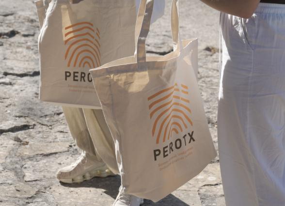 Bolsas de tela promocionales de Perotx con logo de líneas ondulantes naranjas, sostenidas por dos participantes de la actividad.