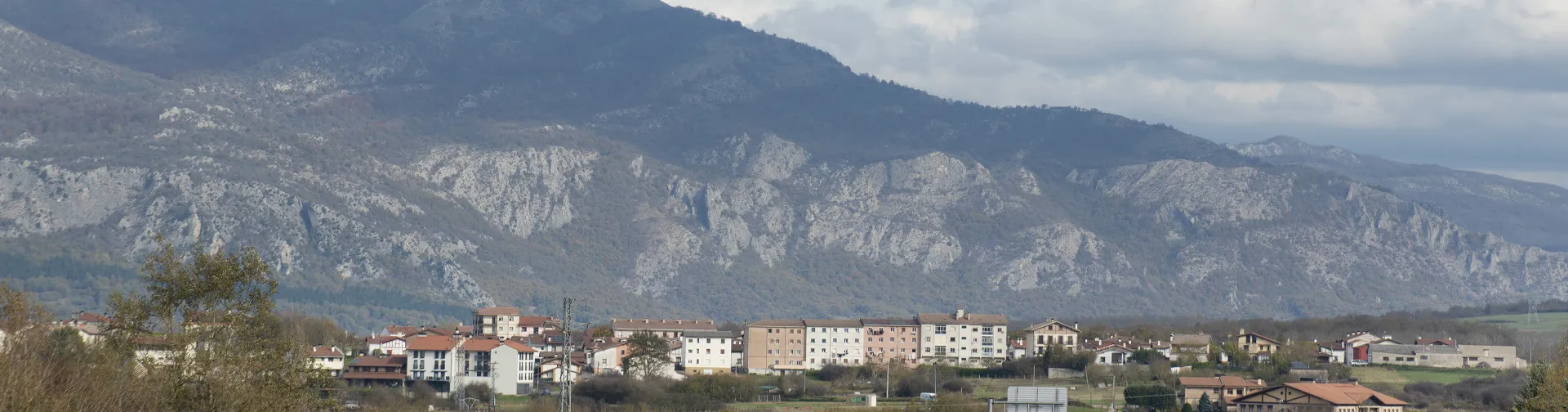 Etxarri Aranaz al pie de la sierra, con montañas al fondo y cielo nuboso en la composición panorámica.