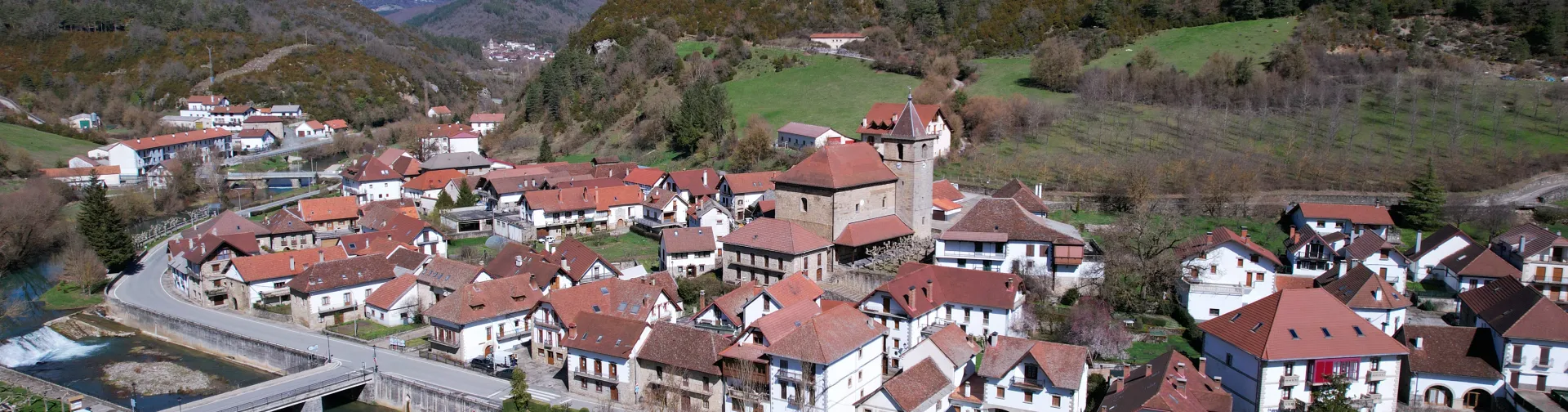 Vista aérea de Ezcaroz mostrando pueblo pirenaico con casas de tejado rojo, iglesia con campanario, valle boscoso y rio.