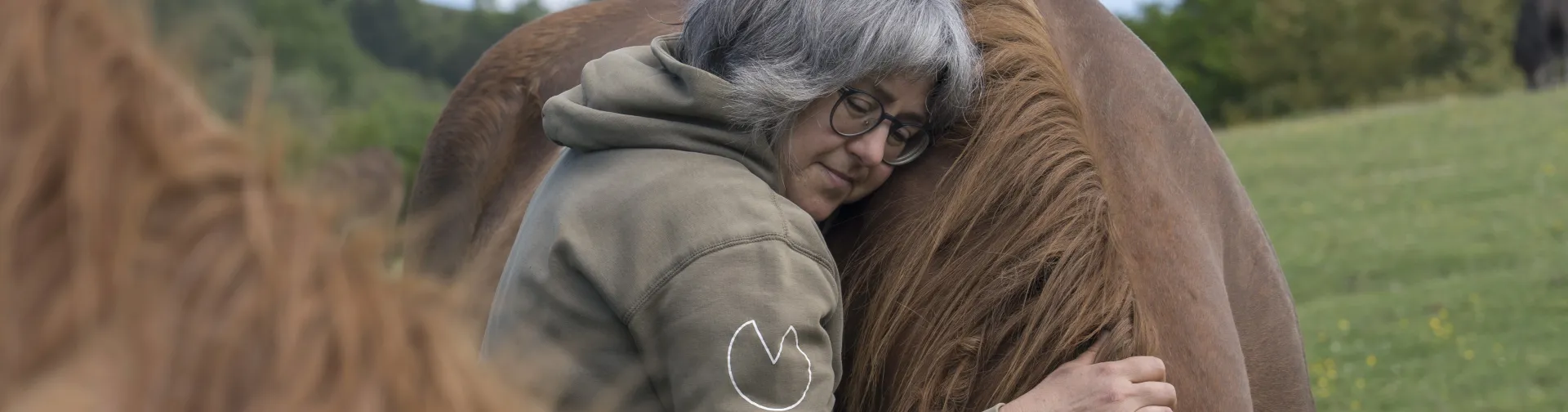 Propietaria de la experiencia de Caballos del bosque abrazando caballo marrón con paisaje de montaña verde al fondo.
