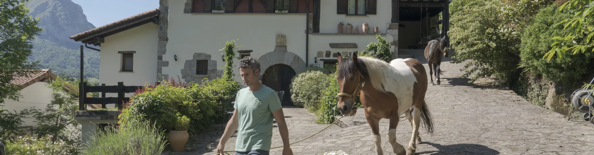 Uno de los propietarios de la experiencia Ekino Aukera paseando caballos con una casa rural tradicional detrás.