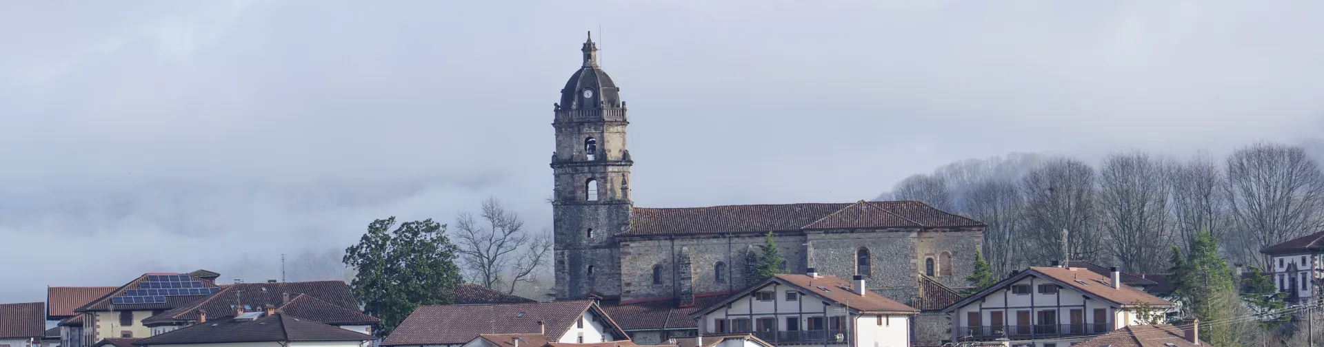 Vista general de Irurita con iglesia de piedra, casas tradicionales blancas y montañas tapadas con las nubes de fondo.