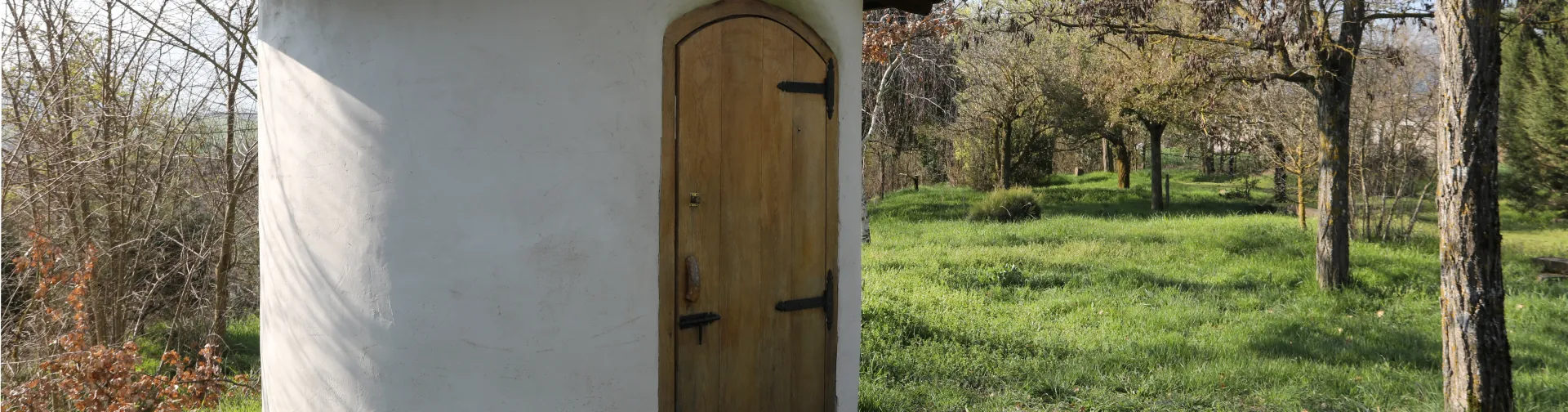 Pequeña caseta de piedra blanca con tejado de barro en Lizoain-Arriasgoiti, rodeada de árboles y prado.