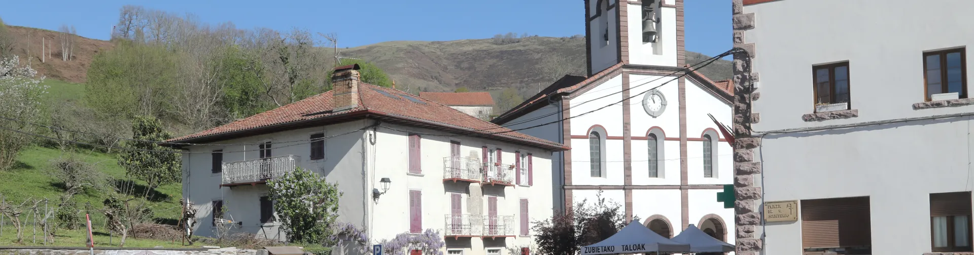 Luzaide-Luzaide plaza, arkitektura tradizionaleko eliza, kanpandorrea eta harrizko eraikinak, bisitariak eta mendiak atzealdean.