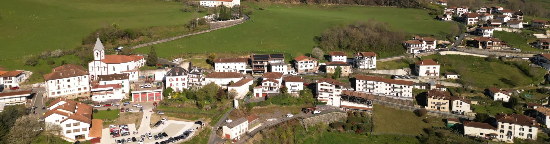 Iglesia blanca con campanario en Luzaide-Valcarlos, rodeada de prados verdes y montañas del Pirineo en valle fluvial.