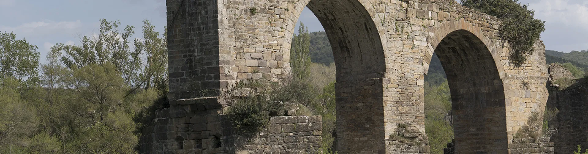 Ruins of a medieval bridge with stone arches over a river in Puente de los Roncaleses, surrounded by vegetation and mountains.