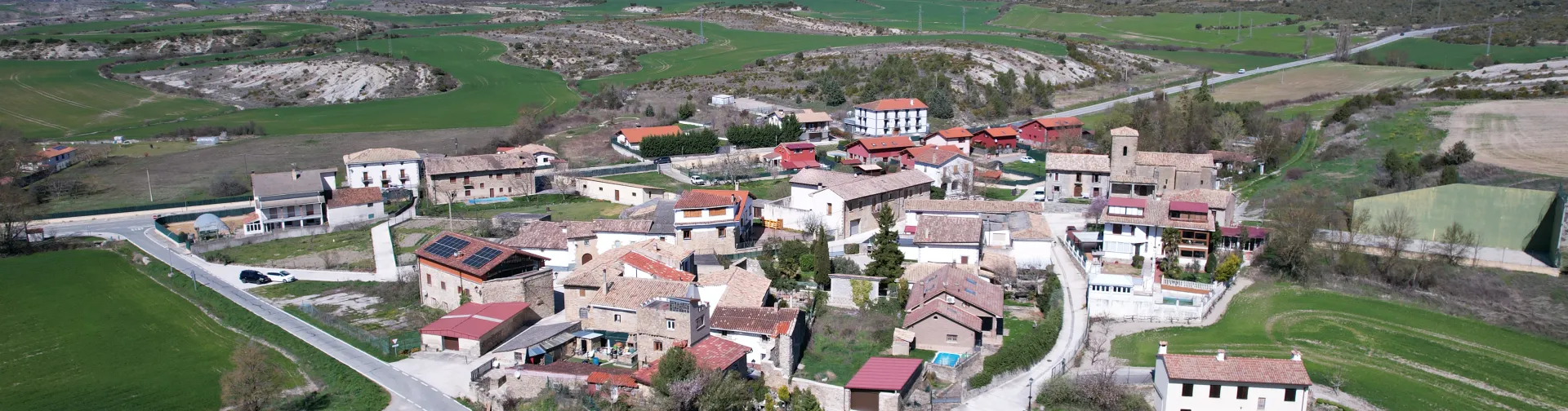 Vista aerea del pueblo de Romanzado con casas dispersas, campos verdes y montana al fondo en el prepirineo.