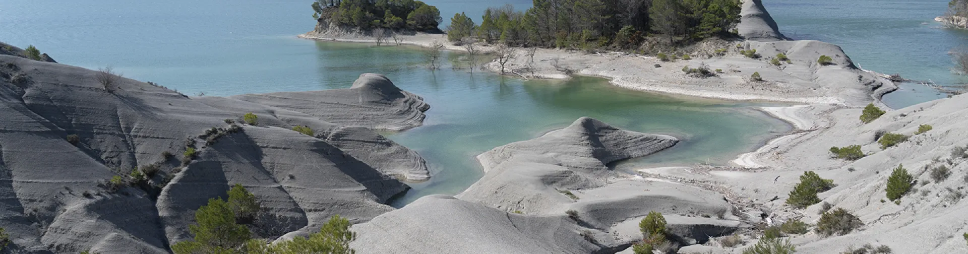 Embalse de Yesa con aguas turquesas, formaciones rocosas grises y vegetación dispersa, montañas al fondo.