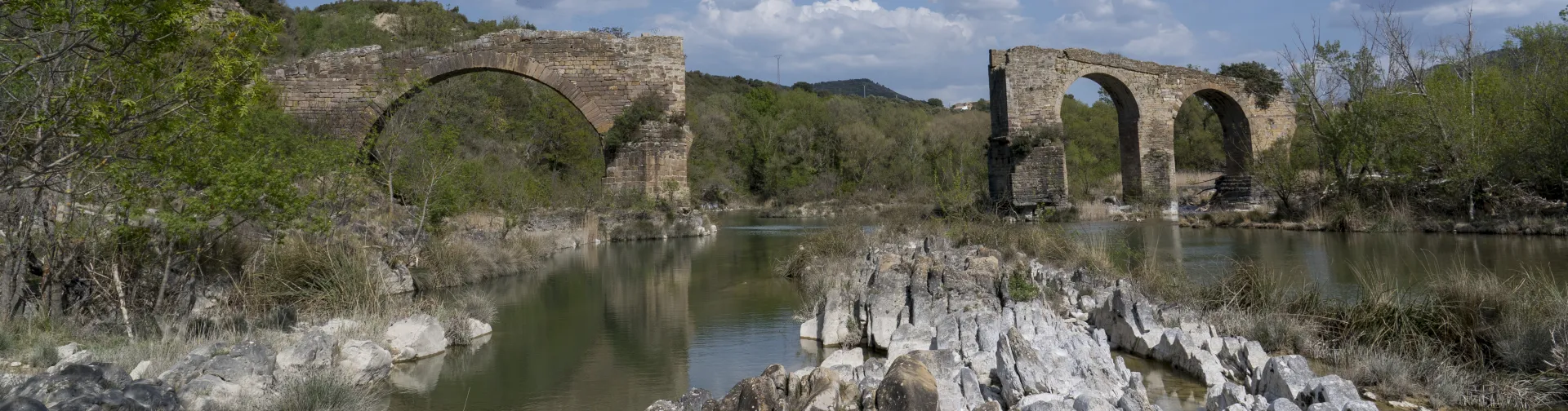 Ruinas de puente medieval con arcos de piedra sobre río en Puente de los Roncaleses, rodeado de vegetación y montaña.