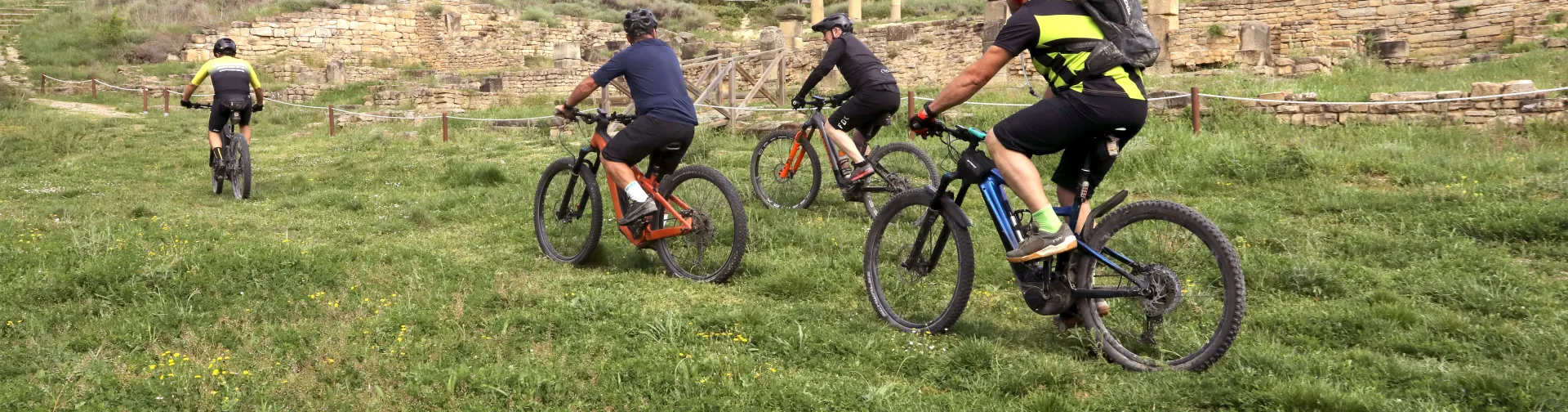 Grupo de ciclistas en prado cerca de ruinas romanas en Territorio Eslava, patrimonio arqueológico al fondo.