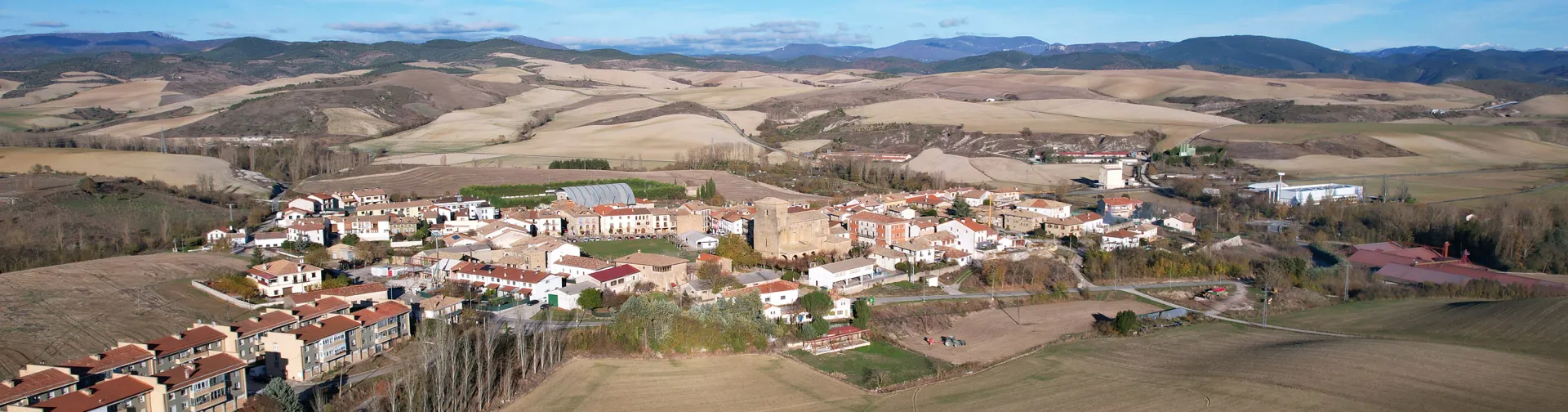 Vista aérea de Urroz-Villa con casas, campos de cultivo y montes al fondo.