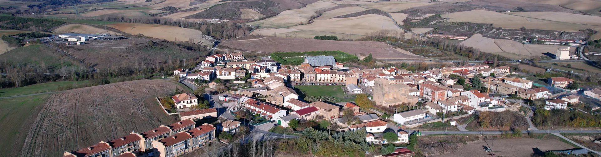 Vista aérea de Urroz-Villa con casas, campos de cultivo y montes al fondo.