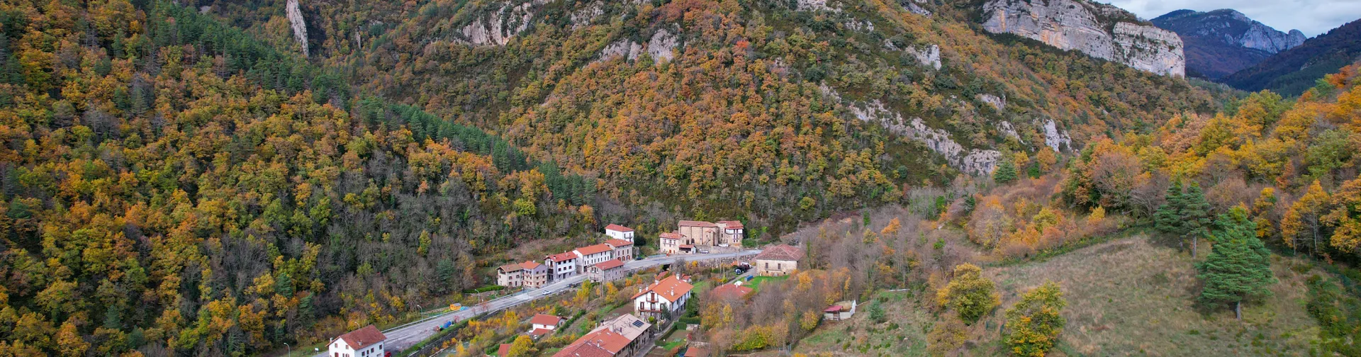Vista aérea de Urzainqui con casas dispersas en valle pirenaico rodeado de bosque en otoño y acantilados de roca caliza.