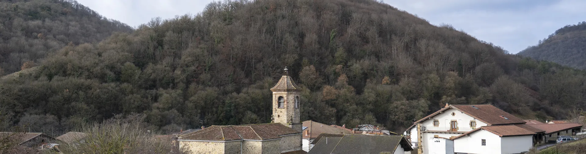 Iglesia con torre campanario de piedra de Anue en entorno de monte verde con casas dispersas.