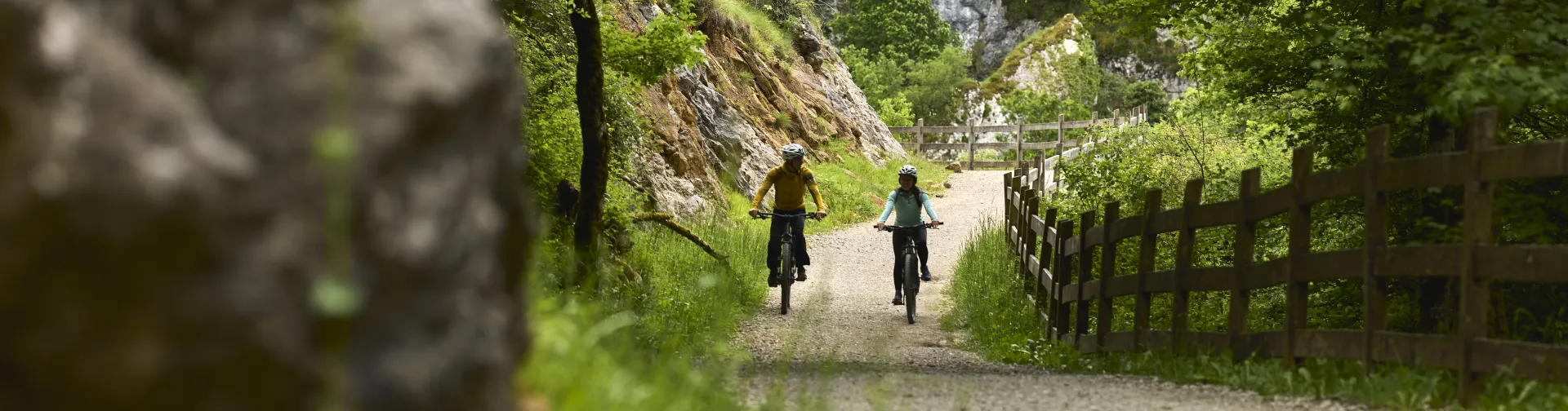 Dos ciclistas en ruta por la Vía Verde del Plazaola pedalean por camino de piedras, a un lado un muro de piedras y vegetación.
