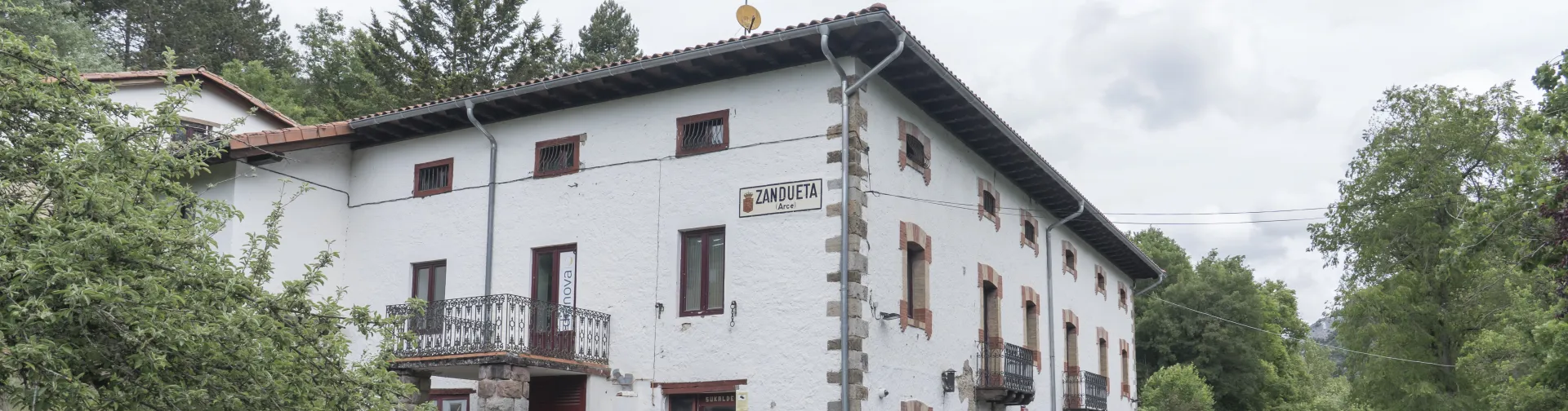 Casa tradicional de Zandueta con fachada blanca y balconada de hierro en entorno boscoso.