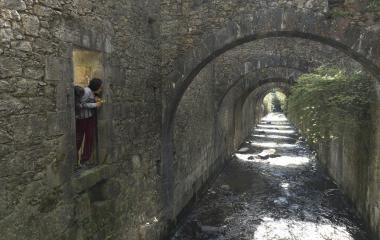 Dos personas se asoman por un balcon en la Fabrica de Armas de Orbaizeta, rio y arcos de piedra al lado.