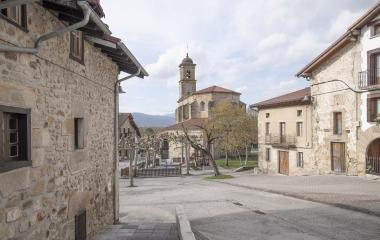Plaza empedrada de Bakaiku con iglesia de piedra al fondo, casas de construcción tradicional y parque ingantil visible.