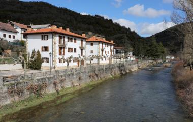 Edificios con tejas naranjas junto al río en Esparza de Salazar, rodeados de bosque de montaña y vegetación.