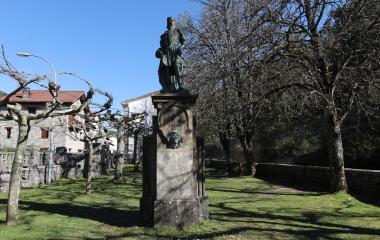 Monumento a personaje histórico en plaza de Garde con casas de piedra y árboles de fondo.