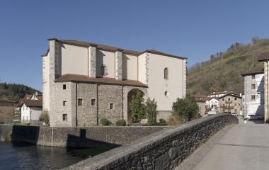 Iglesia de piedra y ladrillo junto al río en Goizueta, con puente de acceso y bosque al fondo.