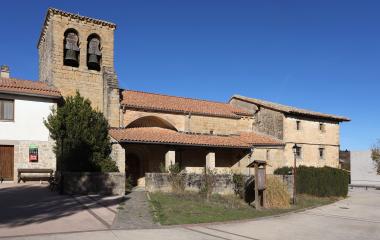 Iglesia de piedra con campanario en Izagaondoa, portico cubierto y tejado de barro bajo cielo azul.