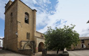 Iglesia de Leache con torre de piedra arenisca, portada y campanario en plaza empedrada.