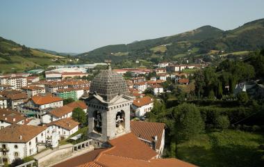 Vista aérea de Leitza con iglesia parroquial de cúpula en primer plano y casas de tejados rojizos entre montes y prados.