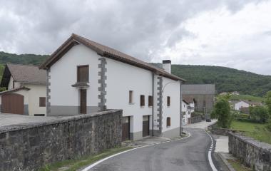 Conjunto de casas tradicionales en el pueblo de Mezkiritz con carretera y muro de piedra en primer plano, monte boscoso al fondo.