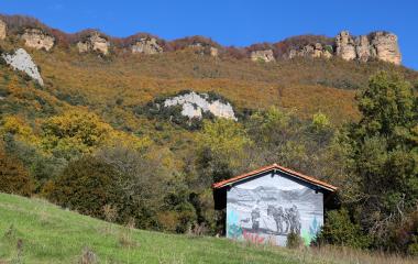 Cabaña con mural en Sierra de Leyre, frente a acantilados calizos y bosque de otoño bajo cielo despejado.