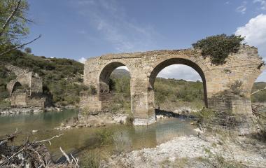 Ruinas de puente medieval con arcos de piedra sobre río en Puente de los Roncaleses, rodeado de vegetación y montaña.