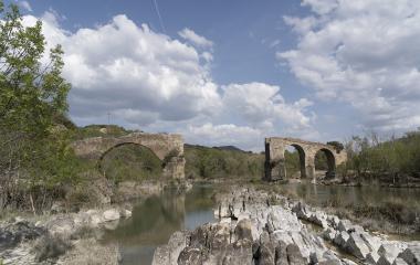 Ruinas de puente medieval con arcos de piedra sobre río en Puente de los Roncaleses, rodeado de vegetación y montaña.