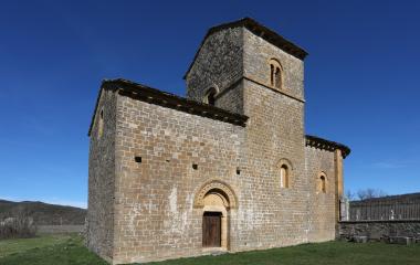 Iglesia románica de piedra en Navascués con torre campanario cuadrada y prado verde al frente bajo cielo despejado.