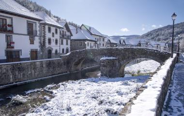 Puente de piedra sobre rio nevado en Ochagavia, con casas blancas y bosque al fondo.