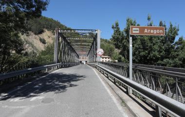 Puente metálico histórico sobre río Aragón en ruta Sendero Urdina, Sangüesa, con cartel indicativo y vegetación riparia.
