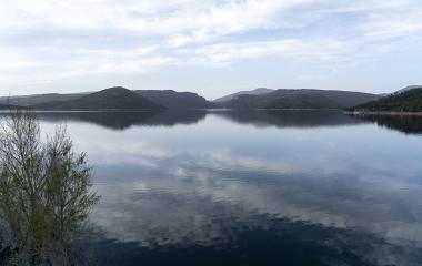 Embalse de Itoitz con montañas boscosas reflejadas en agua tranquila, vegetación en primer plano, cielo nublado.