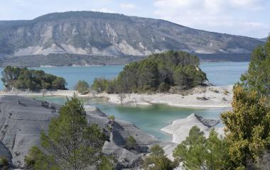 Embalse de Yesa con aguas turquesas, formaciones rocosas grises y vegetación dispersa, montañas al fondo.